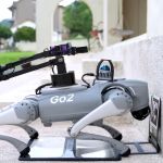 Der vierbeinige Unitree Go2 Roboter steht vor einer Steintreppe, ausgestattet mit einem montierten Roboterarm und Sensorikmodul, während er eine Markierung am Boden scannt.